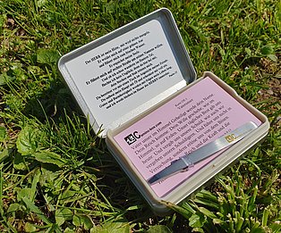 Eine geöffnete Camino Box auf grünem Rasen, der Inhalt enthält neben den Karten auch eine Pinzette